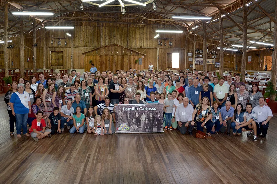 Group photograph from the 3rd Carlesso Family reunion in Chapecó, with around 170 people gathered in a timber hall around the family banner.